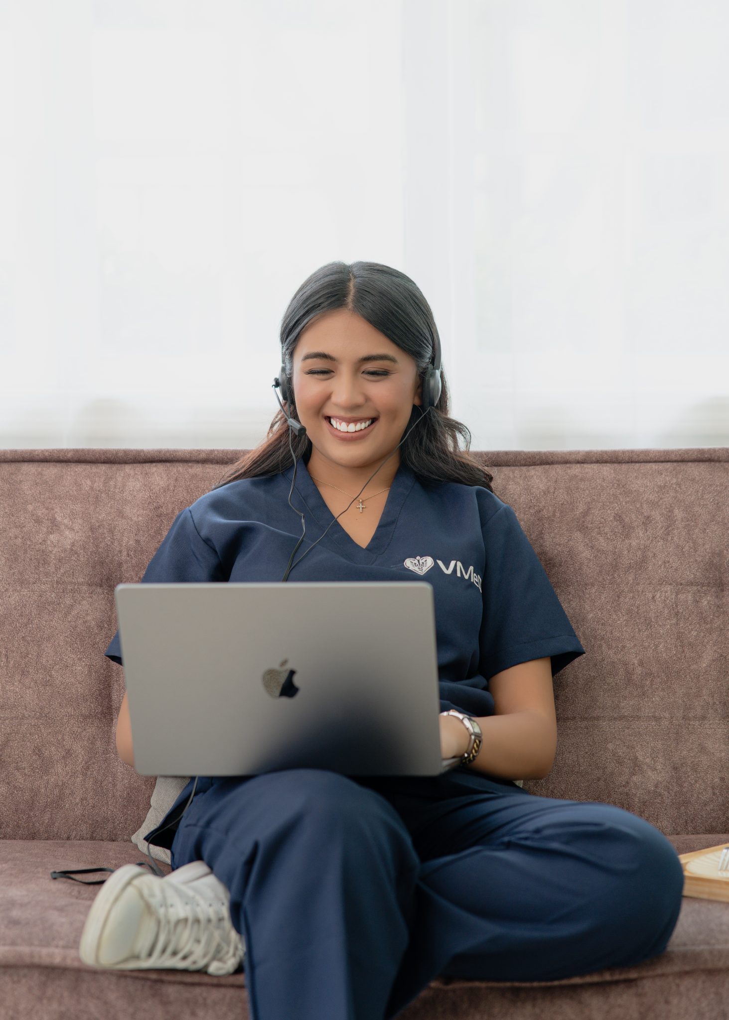 A virtual medical receptionist is sitting on a couch, talking to her client via laptop.
