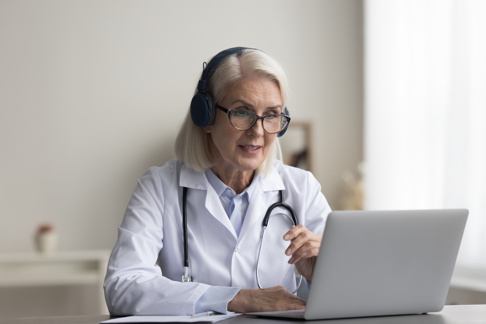 Older female virtual medical scribe specialist sit at desk with modern laptop take part in videocall event, talk to patient, discuss treatment plan.