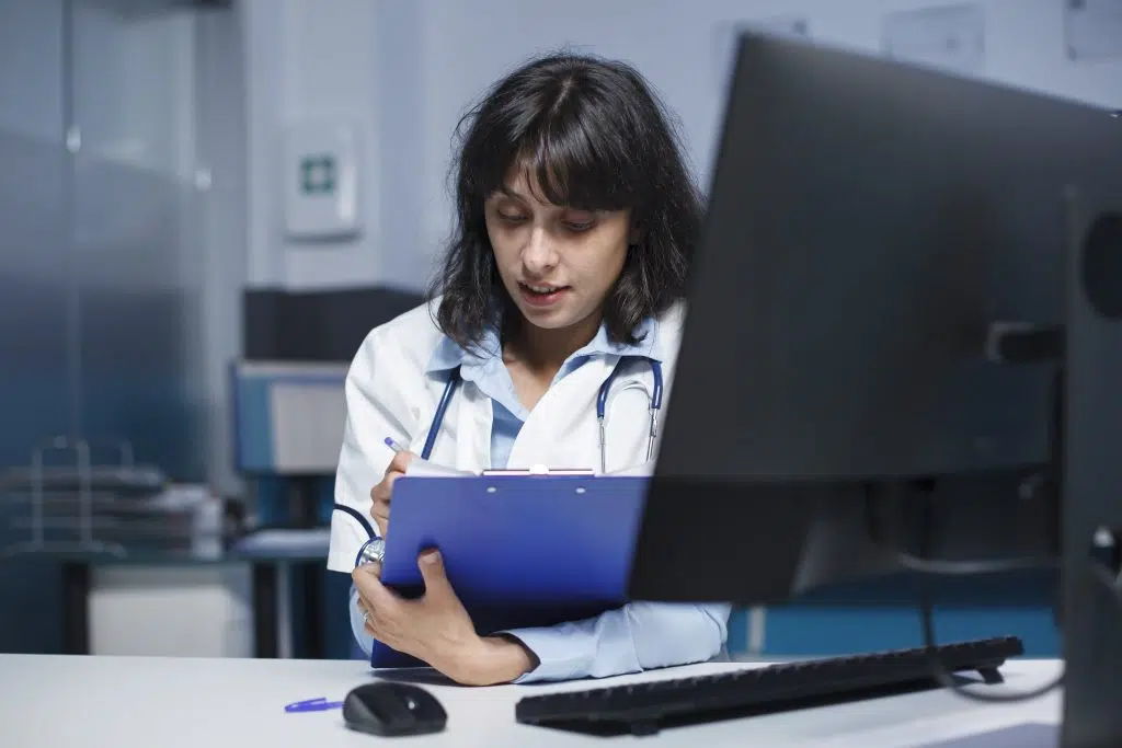 female doctor infront of her screen writing on the clipboard, virtual medical scribe positions