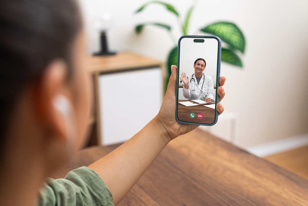A female medical scribe greets a patient through a smartphone video call from home.