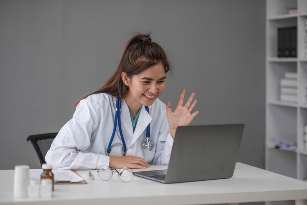A virtual assistant in a white medical uniform with a stethoscope is using a laptop for a video call with a patient, demonstrating essential medical virtual assistant skills.