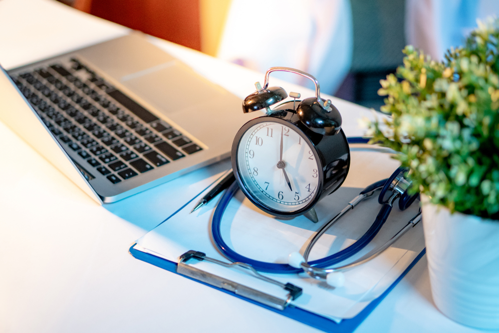 A virtual medical scribe's workspace includes a stethoscope, clipboard, pen, table clock, and laptop on the desk.