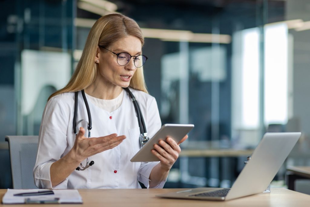 An innovative medical receptionist is conducting a video call with a patient using a tablet and laptop in an office.