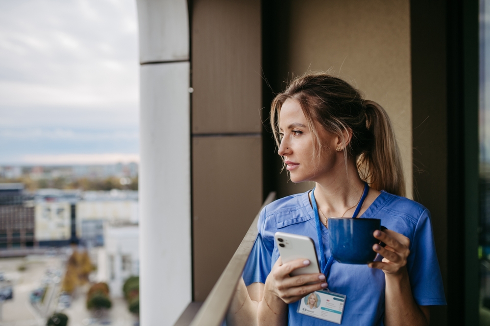 Female medical scribe enjoying cup of coffee at home after work, standing by window. Morning tea before work. Work-life balance for healthcare worker.