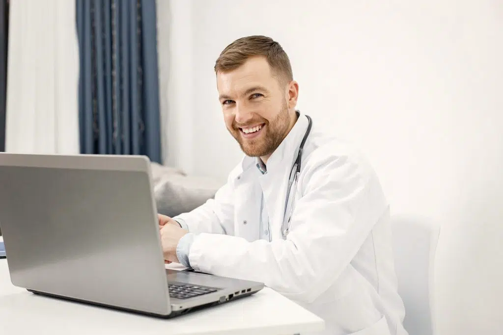 A doctor smiles while using his laptop, representing the best virtual medical scribes in healthcare