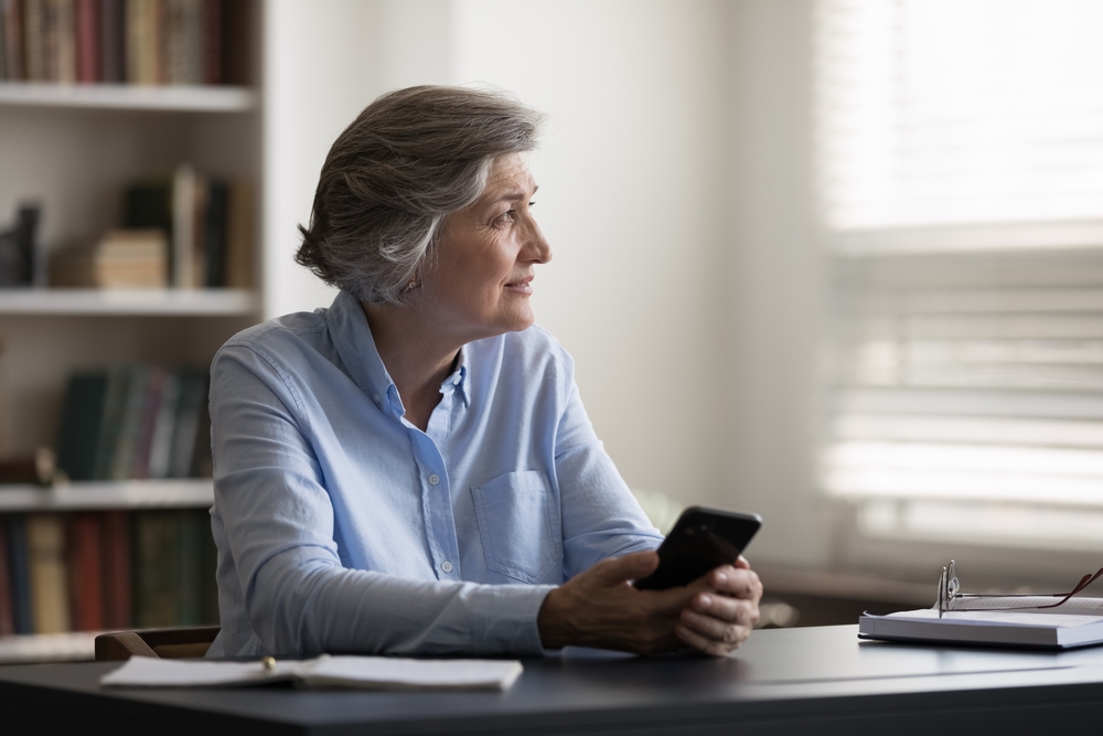 virtual medical receptionist, mature middle aged woman sitting at table with cellphone in hands, looking in distance.