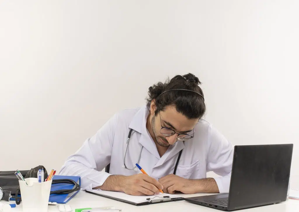 A male doctor writes notes while using his laptop, reflecting on how much a virtual medical scribe costs.