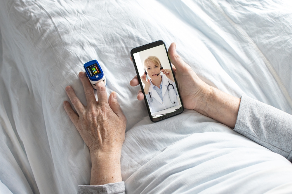 An elderly woman measures her pulse and oxygen levels with an oximeter during an online consultation with a doctor while sitting in bed under a white blanket.
