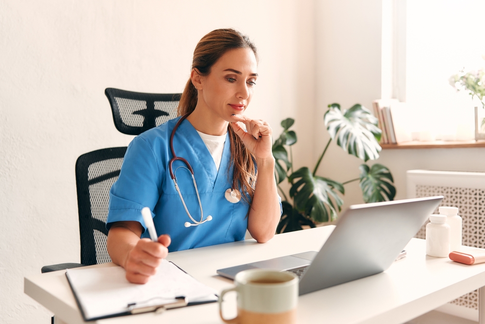 Female medical scribe at home in medical uniform with stethoscope sitting at table with laptop talking on video call in living room.