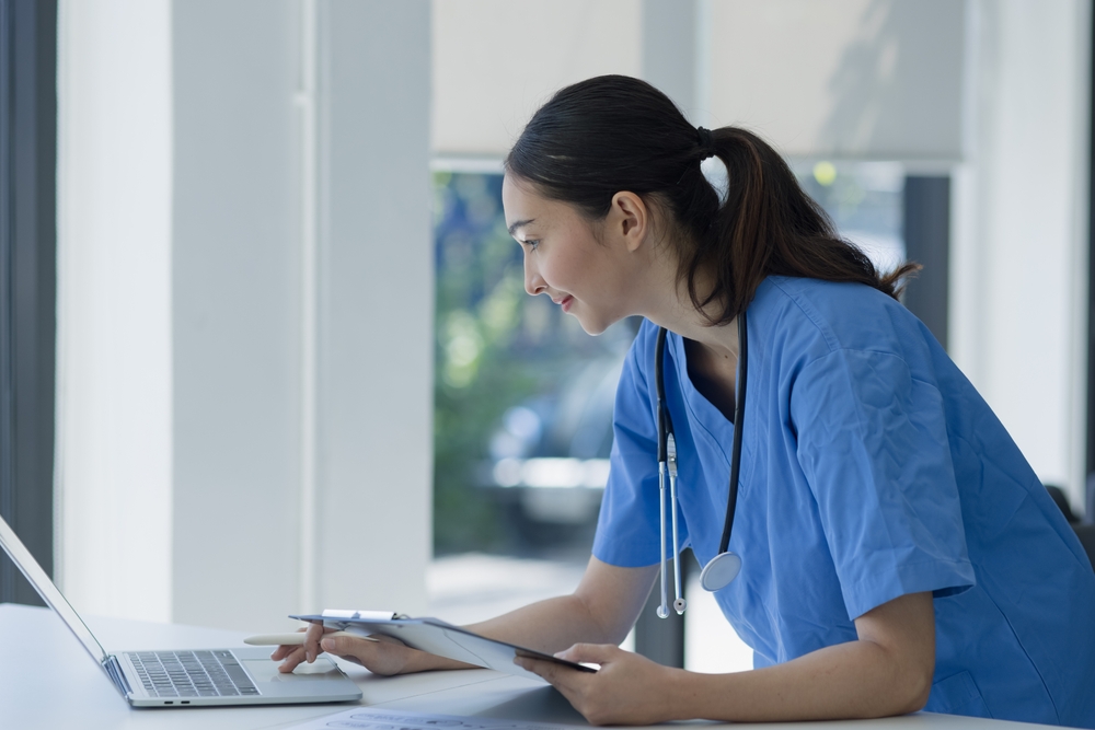 Female medical scribe sitting at a desk with a laptop and clipboard, exploring medical scribe virtual jobs.