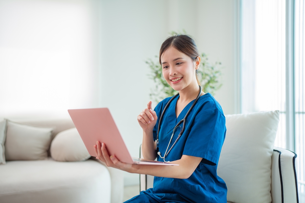  Medical scribe specialist wearing medical scrubs with a stethoscope using a laptop talking to her patient while sitting on sofa.