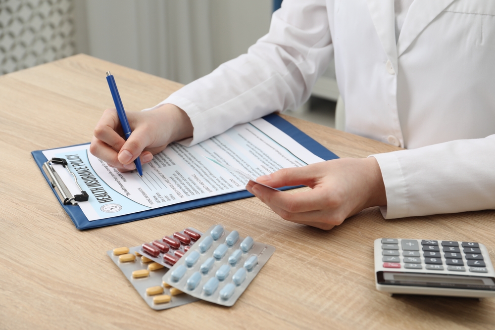 A doctor signing a form on a table with medicine tablets and a calculator