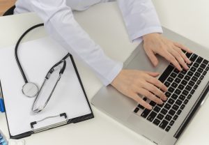 cropped shot of female doctor hands working with laptop at desk with medical tools and clipboard, accurate medical transcription services 