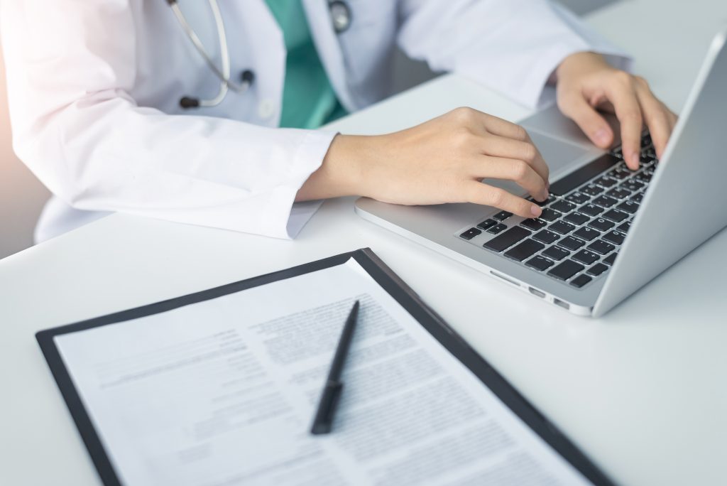 Concentrated Asian woman Doctor hands using laptop computer and working on checklist paper symptoms of patients on a clipboard in the medical room of the hospital. accurate medical transcription services