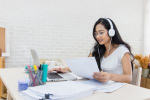 A young Asian woman working online from home. She uses a headset video conference calling on laptop computer talk by webcam. She avoids the outbreak of coronavirus. The Internet is everything., academic transcription services for lecturers