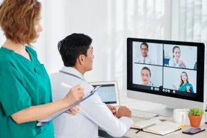 Chief physician having online conference with colleagues when his assistant taking notes in documents, remote healthcare communication team