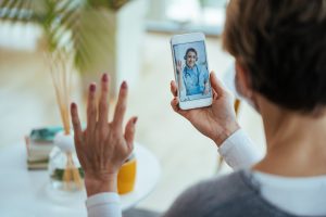 Close-up of woman greeting her doctor while using smart phone and having video call. Focus is on female doctor on touchscreen, international patient scheduling services