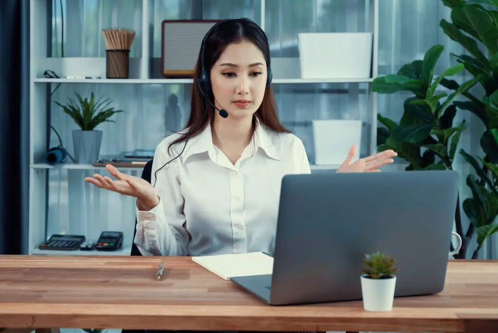 Asian call center with headset and microphone working on her laptop. Female operator provide exceptional customer service. Supportive call center agent helping customer on inquiry. Enthusiastic, virtual medical front desk staff