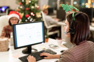 Female operator in xmas headband and headset sitting in front of computer screen and shopping online by workplace, thanksgiving efficiency receptionist