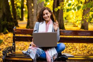 Happy woman talking on line in a video conference with a laptop sitting on a bench in the street, autumn efficiency receptionist
