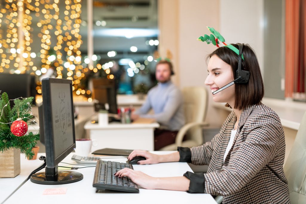 Happy young operator in xmas headband and headset looking at computer screen while shopping online by workplace, thanksgiving efficiency receptionist