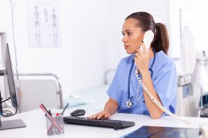 Medical nurse talking with patient on the phone about diagnosis. Health care physician sitting at desk using computer in modern clinic looking at monitor, international medical call handling