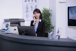 Portrait of smiling hospital receptionist answering call from patient to make an appointment for clinical consult at private clinic. Professional looking woman working at front desk using telephone, virtual medical front desk staff