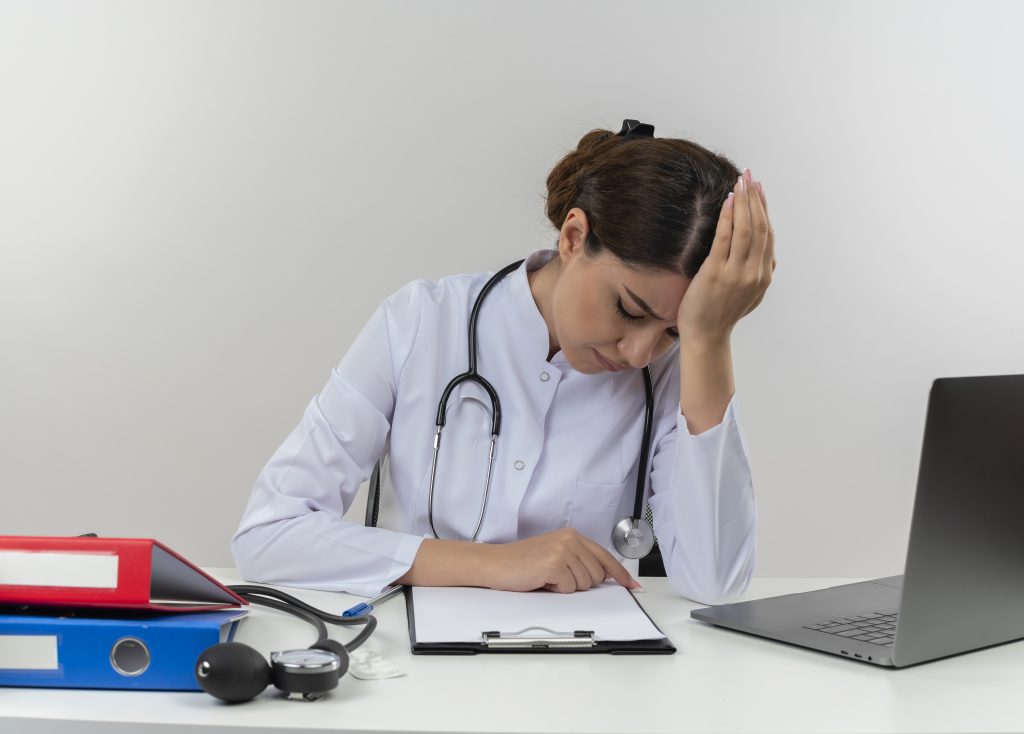 tired young female doctor wearing medical robe with stethoscope sitting at desk work on computer with medical tools lowered head on isolated white background with copy space, healthcare challenges receptionist