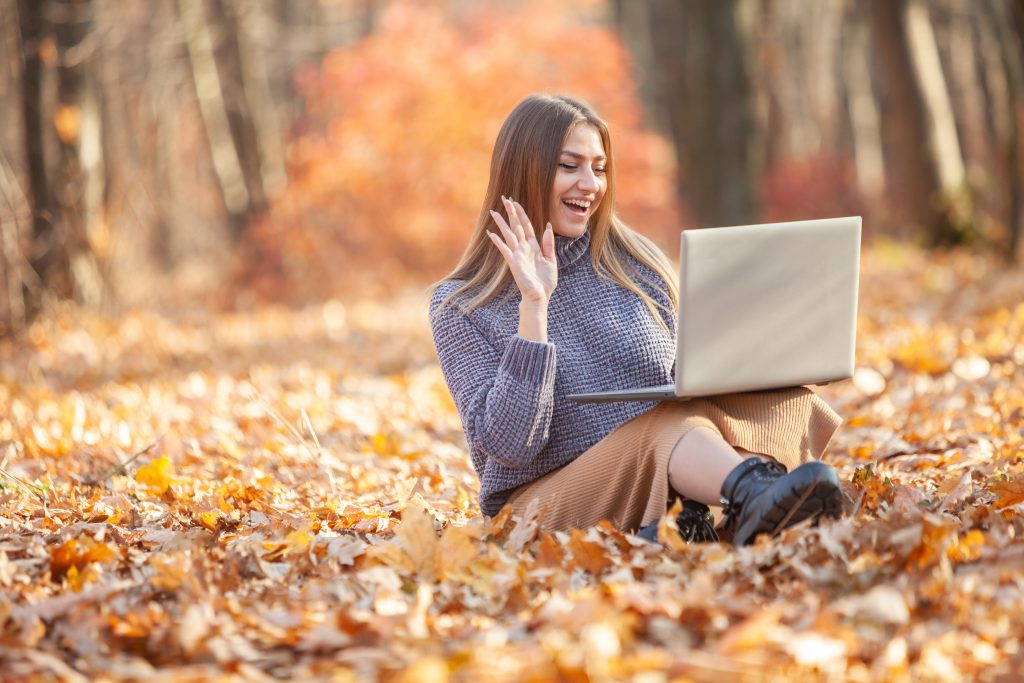 Young female freelancer or student with a laptop waving at the screen while sitting on the fallen leaves in the forest. Remote work or education. Video call, blogger and blogging concept, halloween service receptionist
