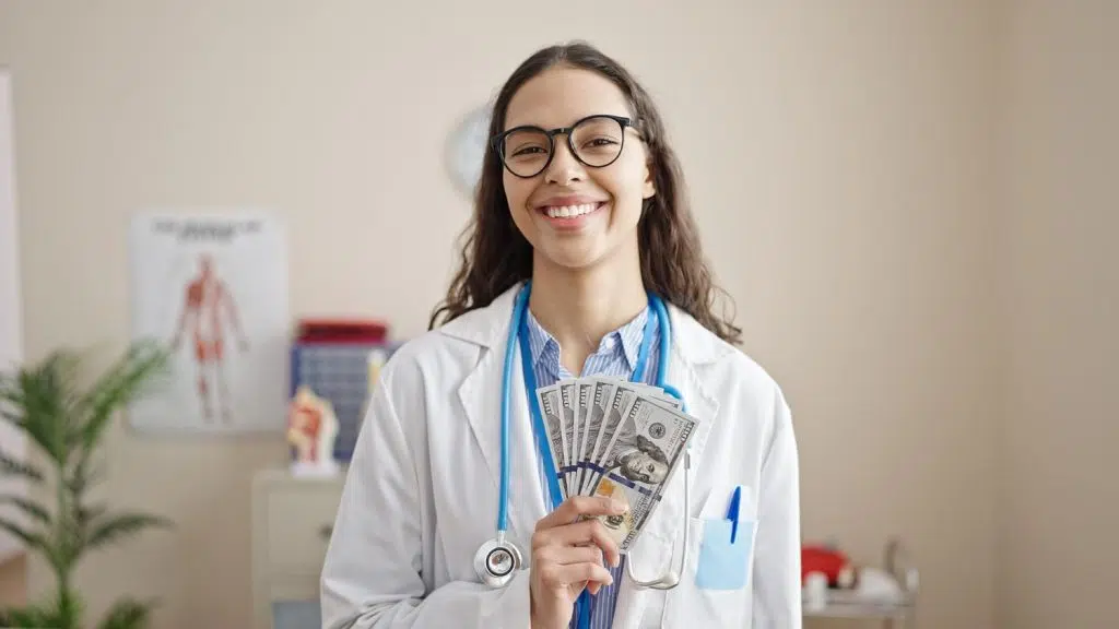 Smiling female doctor holding money in a clinic representing patient care coordinator salary