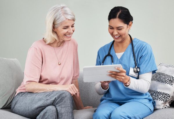 Care coordinator showing mature patient medical results and online healthcare report on tablet while sitting on sofa.