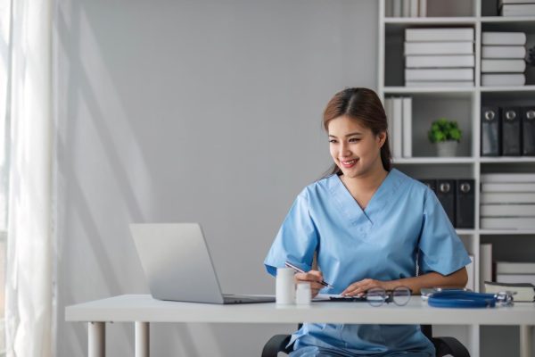 Clinical virtual assistant working on a laptop and writing on a clipboard at a hospital office desk.