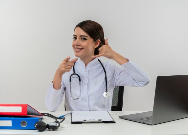 Female healthcare professional in medical robe with stethoscope, sitting at desk using computer, medical tools nearby, making phone call gesture with hand.