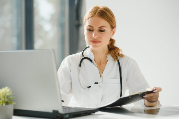 Medical receptionist writing on clipboard, and working on her laptop.