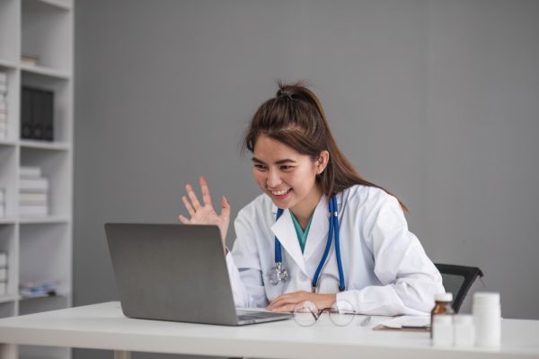 Virtual medical assistant with stethoscope waving at patient on laptop screen.