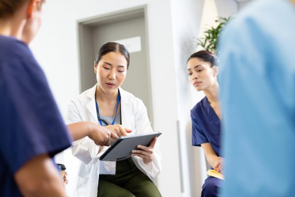 female doctor with tablet sitting in discussion with diverse female colleagues at hospital.