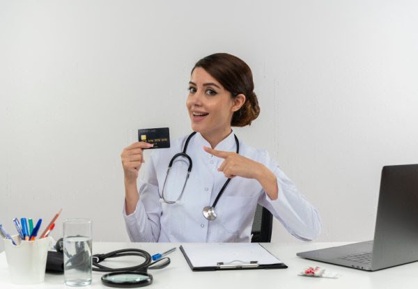 Medical billing assistant in medical robe with stethoscope, sitting at desk with medical tools and laptop, holding and pointing at credit card.